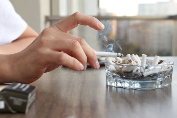 A person holding a lit cigarette over an ashtray filled with old cigarette butts.